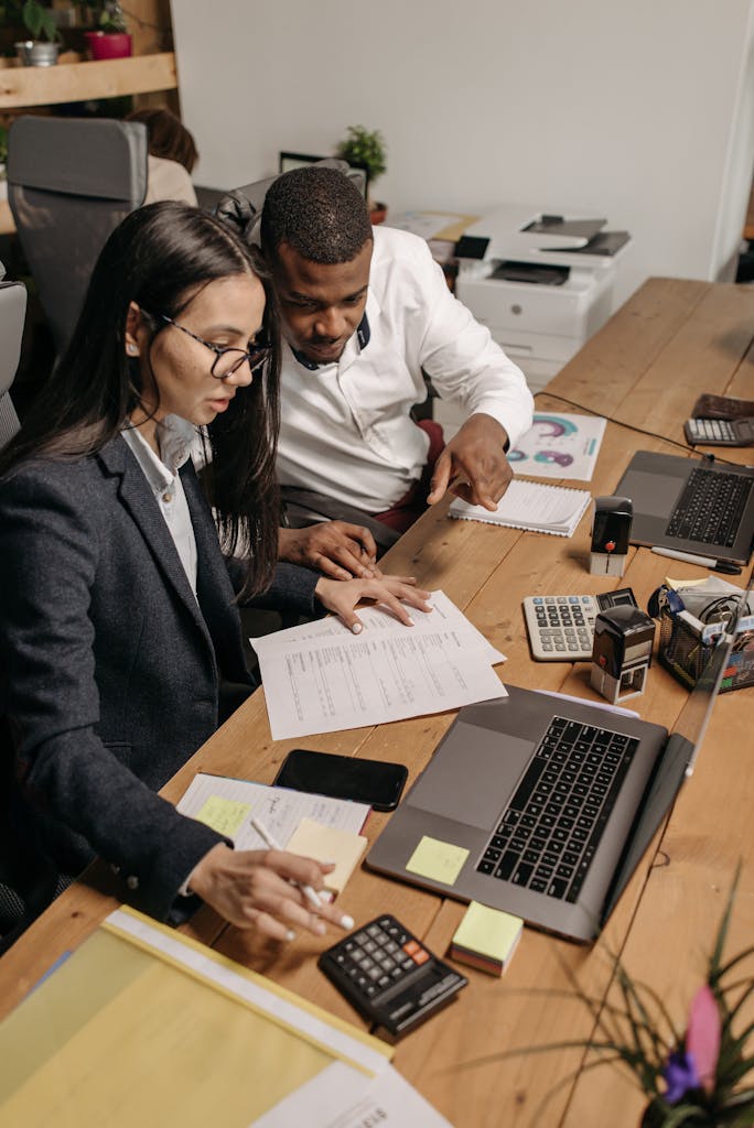 Two employees working together on financial documents with laptops and calculators in an office setting.